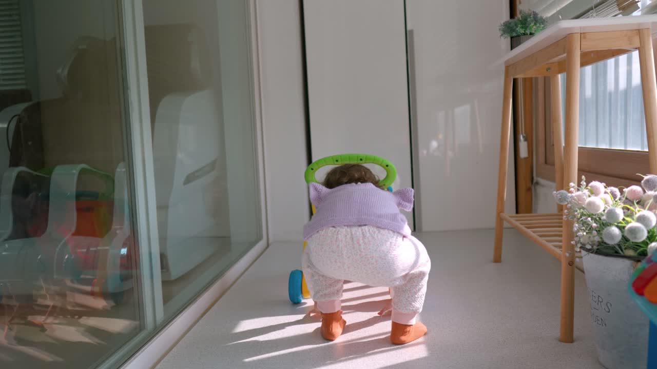 маленькая девочка учится ходить с поддержкой сидячих ходунков на балконе - вид сзади замедленного действия