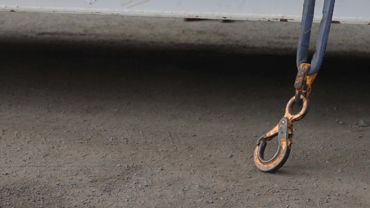 A worker in an orange shirt steps away from a winch attached to an industrial crane hook, showcasing industrial operations and machinery.
