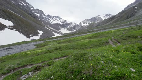 Fast-aerial-drone-footage-flying-close-over-an-alpine-meadow-before-tilting-up-to-reveal-a-rugged,-glacially-carved,-snow-covered-mountain-landscape-and-a-remote-hiking-trail-in-Switzerland