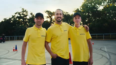 Fröhliches-Und-Freundliches-Fahrlehrertrio-In-Einer-Fahrschule.-Drei-Typen-In-Gelben-T-Shirts-Posieren-Vor-Motorrädern