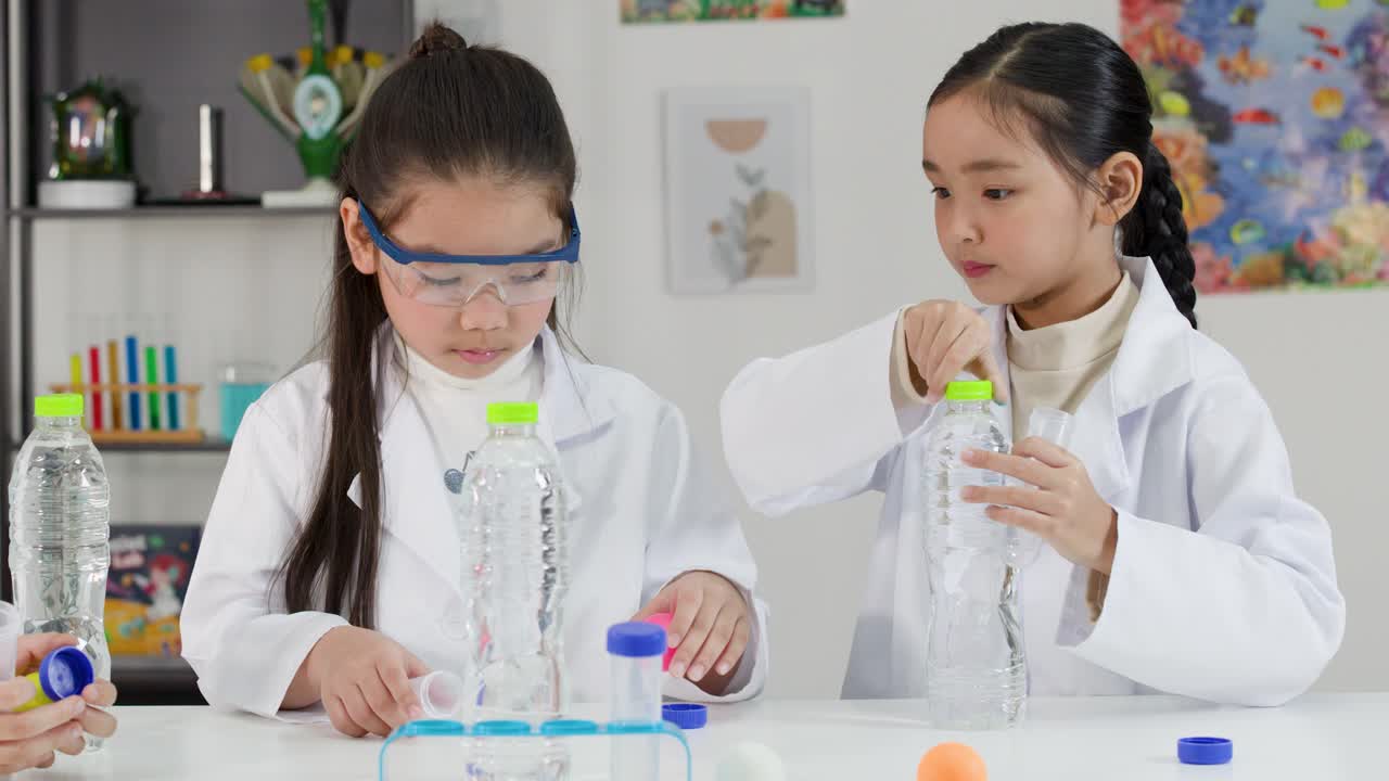 Dua gadis Asia mengenakan jas lab melakukan percobaan fisika menggunakan botol plastik dan bola.