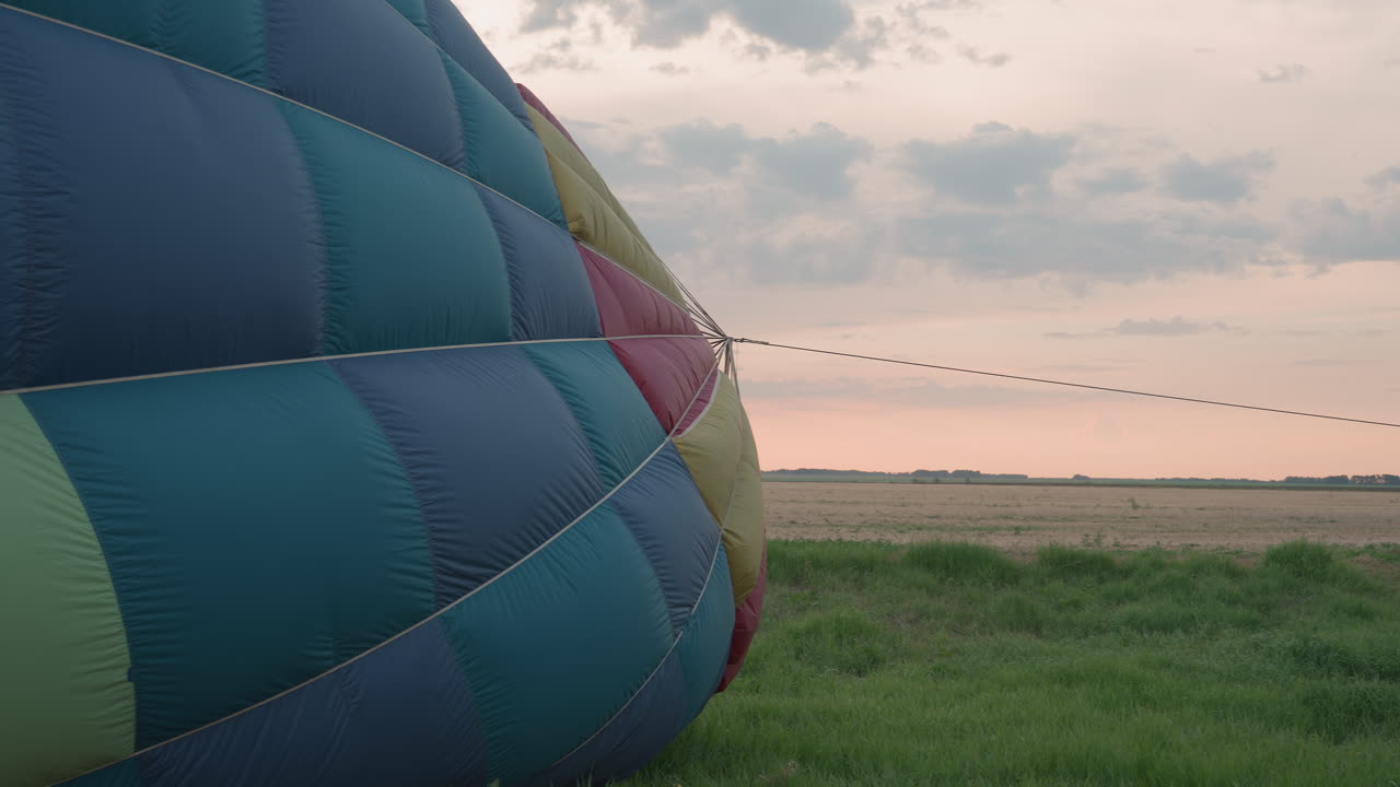 young man in tall grass field holding rope to secure hot air balloon envelope before launch amid sunrise sky soft clouds and open farm land landscape with calm early morning atmosphere