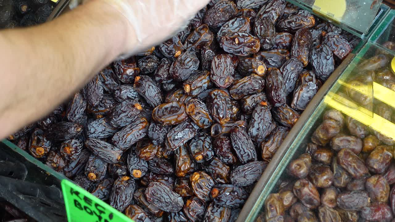 A person's hand wearing gloves picks up dates from a bin at a market