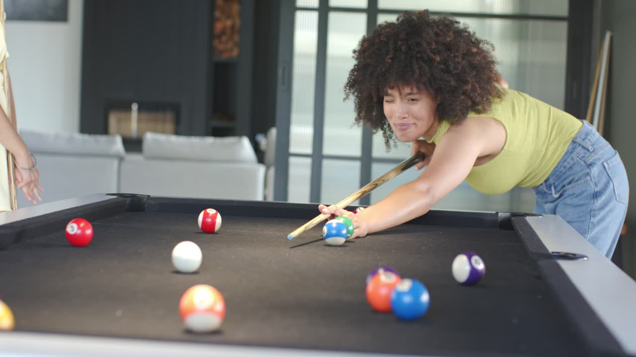 African American woman playing pool with friend at home, taking turn lining up cue to score