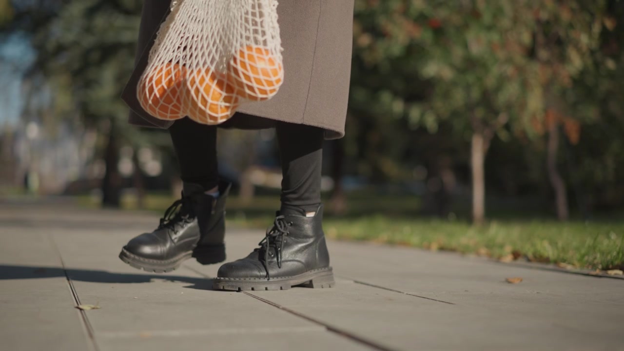 Primo piano di una donna che cammina sul marciapiede tenendo una borsa a rete arancione con frutta. I suoi stivali neri e il cappotto marrone sono visibili mentre la donna si muove attraverso la scena portando borse della spesa riutilizzabili. Sfondo.
