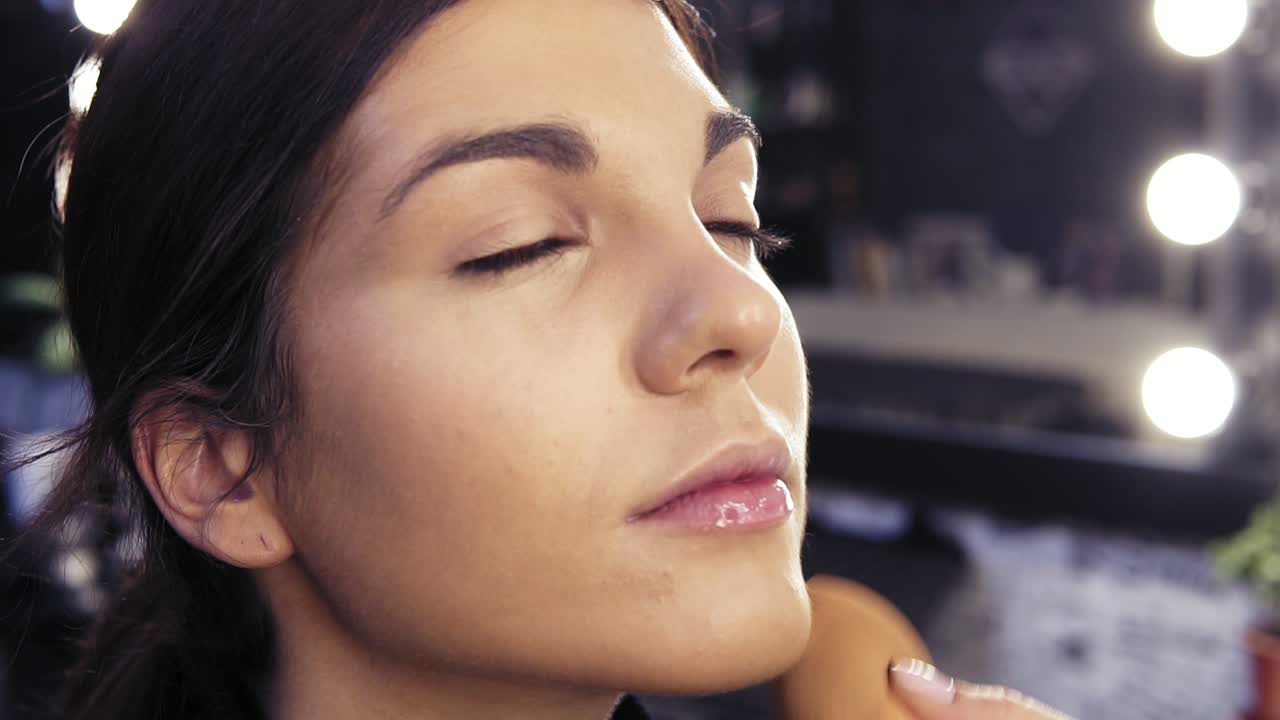 Make up artist's hand applying foundation on previously prepared face of an attractive brunette girl in a beauty salon or studio.