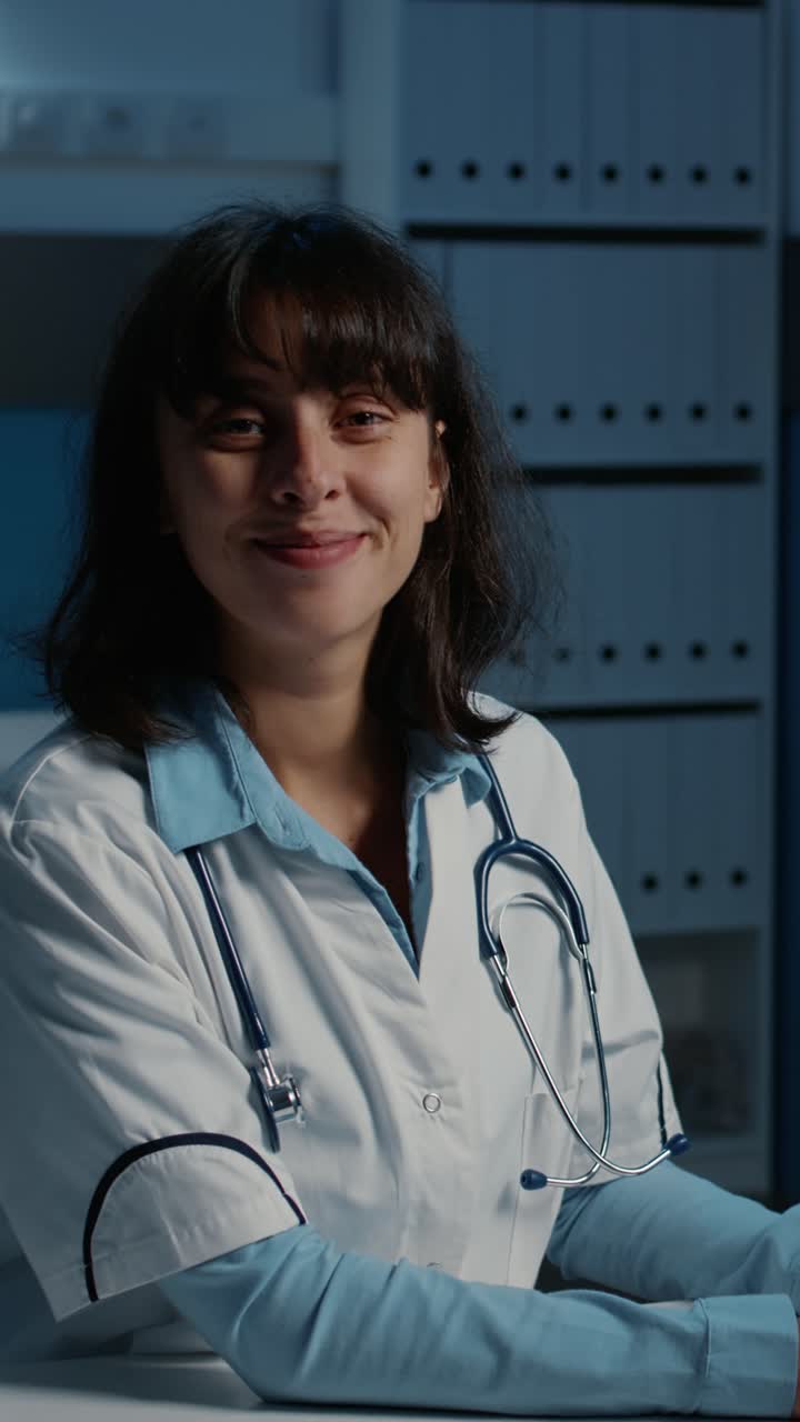 Vídeo vertical: Retrato de un médico sonriente sentado en su escritorio escribiendo información médica en el ordenador
