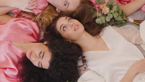 top view of group of multiethnic female friends and bride dressed in pink and white silk nightdresses holding hands with bouquets while laying on floor