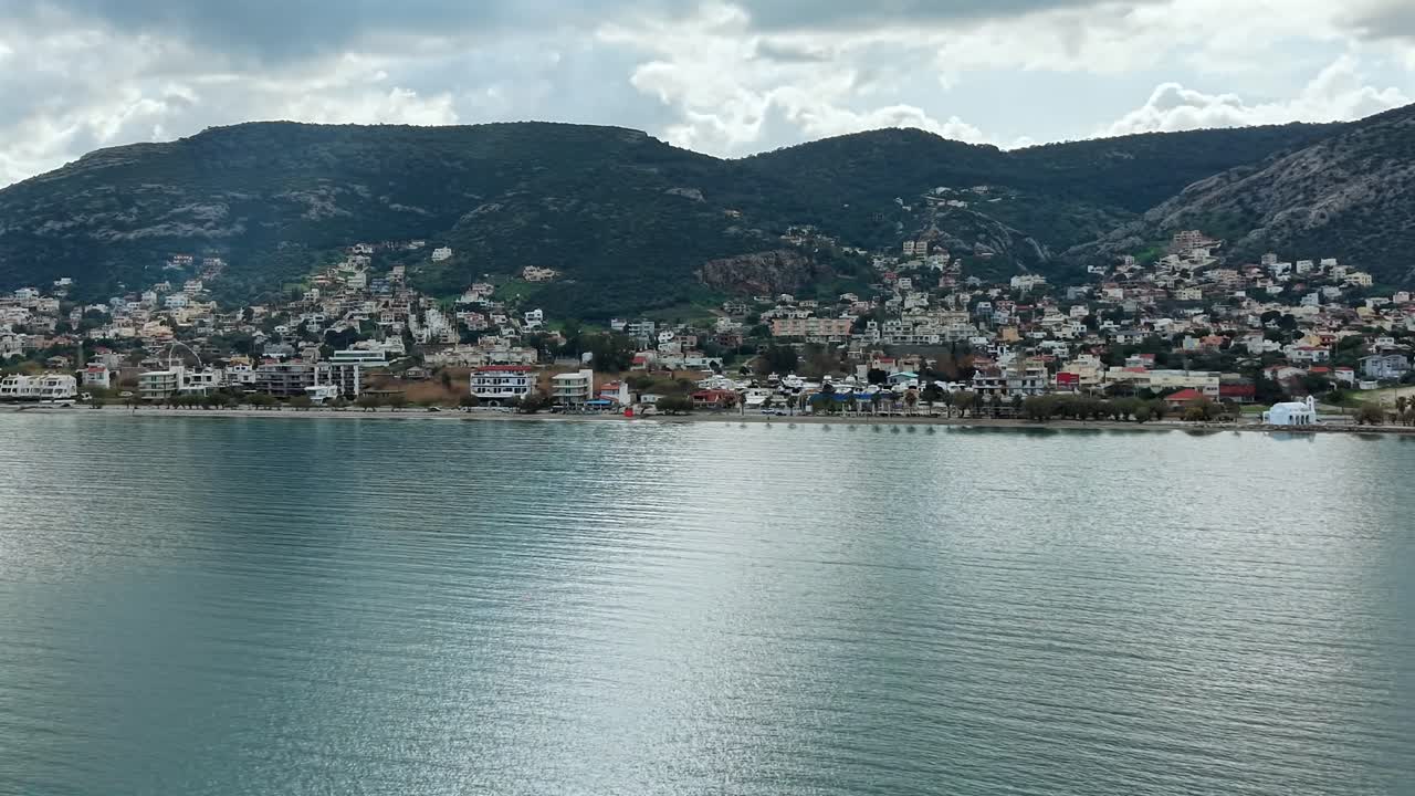 Drone, Porto Rafti kıyısına paralel olarak turkuaz Ege Denizi üzerinde 110 m yükseklikte seyrediyor. Gür yeşil tepeler kasabanın arkasında yükseliyor; beyaz bulutlarla aydınlık gökyüzü. Sakin sularda parıldayan güneş ışığı, Ocak 2026