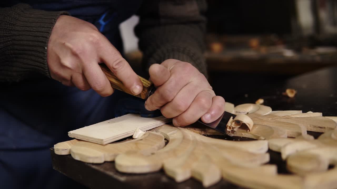 Slow motion of carpenter working on a wooden in his workshop on the table, preparing a detail of wooden product, a part of future furniture. Close up footage of a man's hands cuts out patterns with a planer