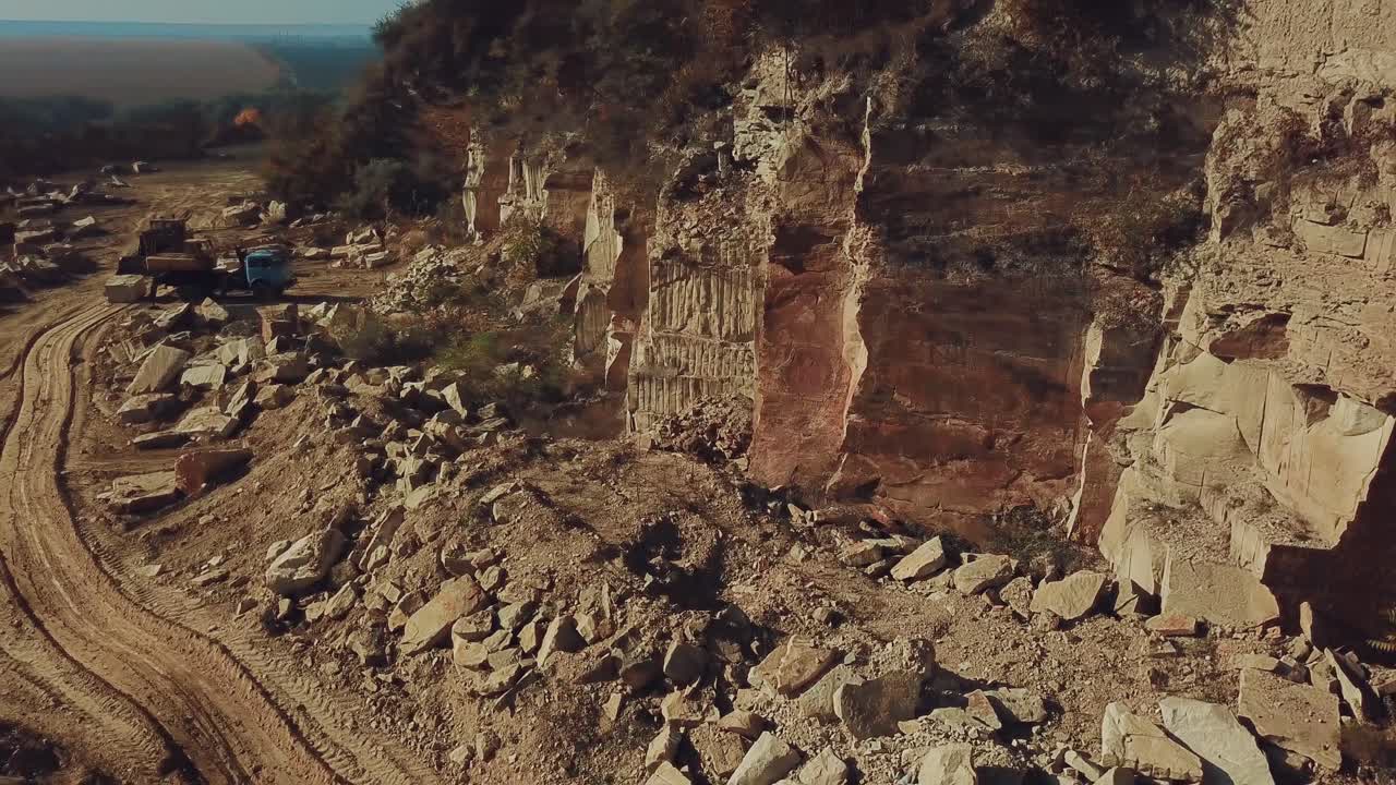A large quarry with stone rocks and trees at the top is located in the countryside near a dirt road. Aerial view.
