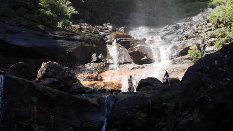 people walking and enjoying a rock pool at the bottom of a waterfall during a sunny day with high contrast