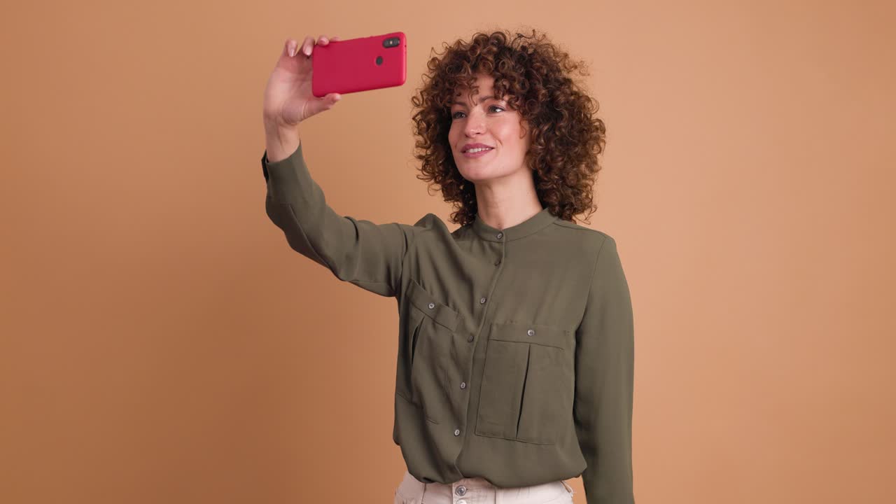 Glad kvinde tager selfies med mobiltelefon på beige baggrund.