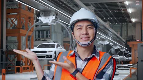 close up de ingeniero masculino asiático con casco de seguridad sonriendo y apuntando a un lado mientras está de pie en la fábrica de automóviles ev