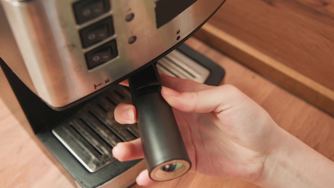 Closeup of hands guiding portafilter into stainless espresso machine on wooden counter, showing careful coffee preparation, manual rotation, secure locking, and warm cafe reflections