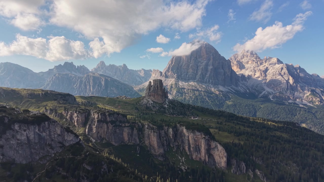 Veduta aerea con drone delle Cinque Torri che si ergono sopra aspre scogliere calcaree e una foresta alpina, con l'imponente massiccio delle Tofane e le cime dolomitiche circostanti