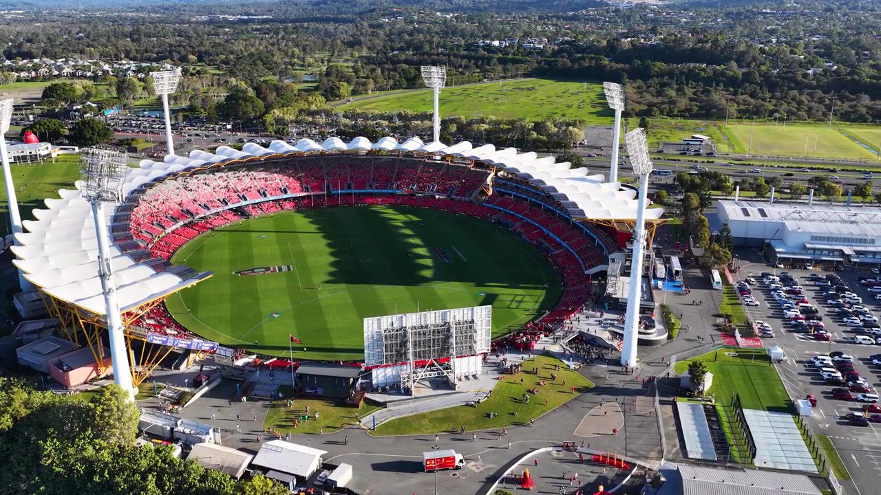 Wide drone shot of a circular sports arena with green grass under bright daylight.