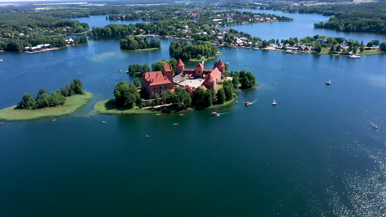 공중: 트라카이 성 주변의 호수를 돌고 있는 보트와 요트가 있는 트라카이 성의 높은 고도 회전 샷, 호수 표면에 태양이 빛을 반사하는 모습
