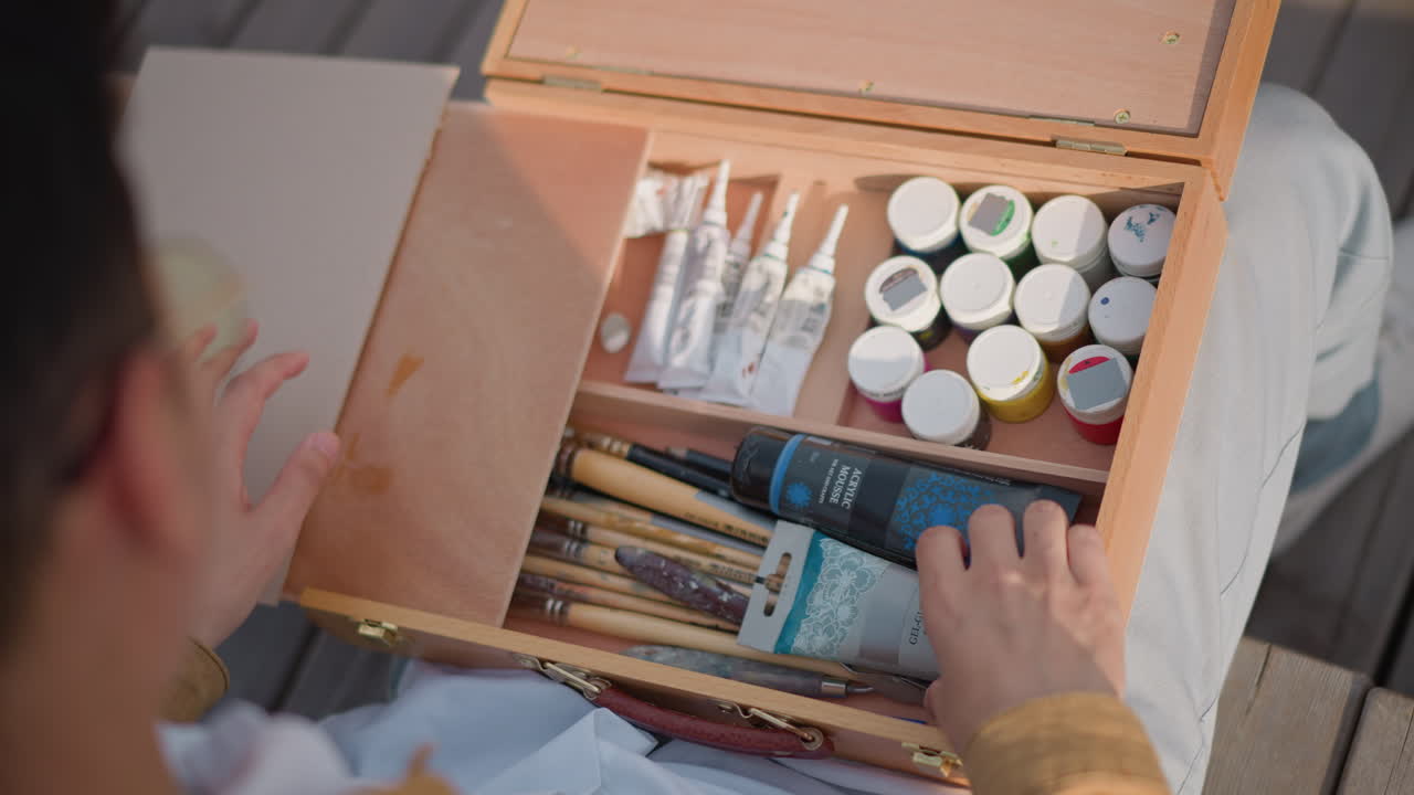 artista apre scatola di legno con pennelli, tubetti e piccoli barattoli, sistema la palette, seleziona un cartone per schizzi di strada, ripresa spontanea sopra la spalla, toni illuminati dal sole, movimenti tattili, vivace ambiente urbano