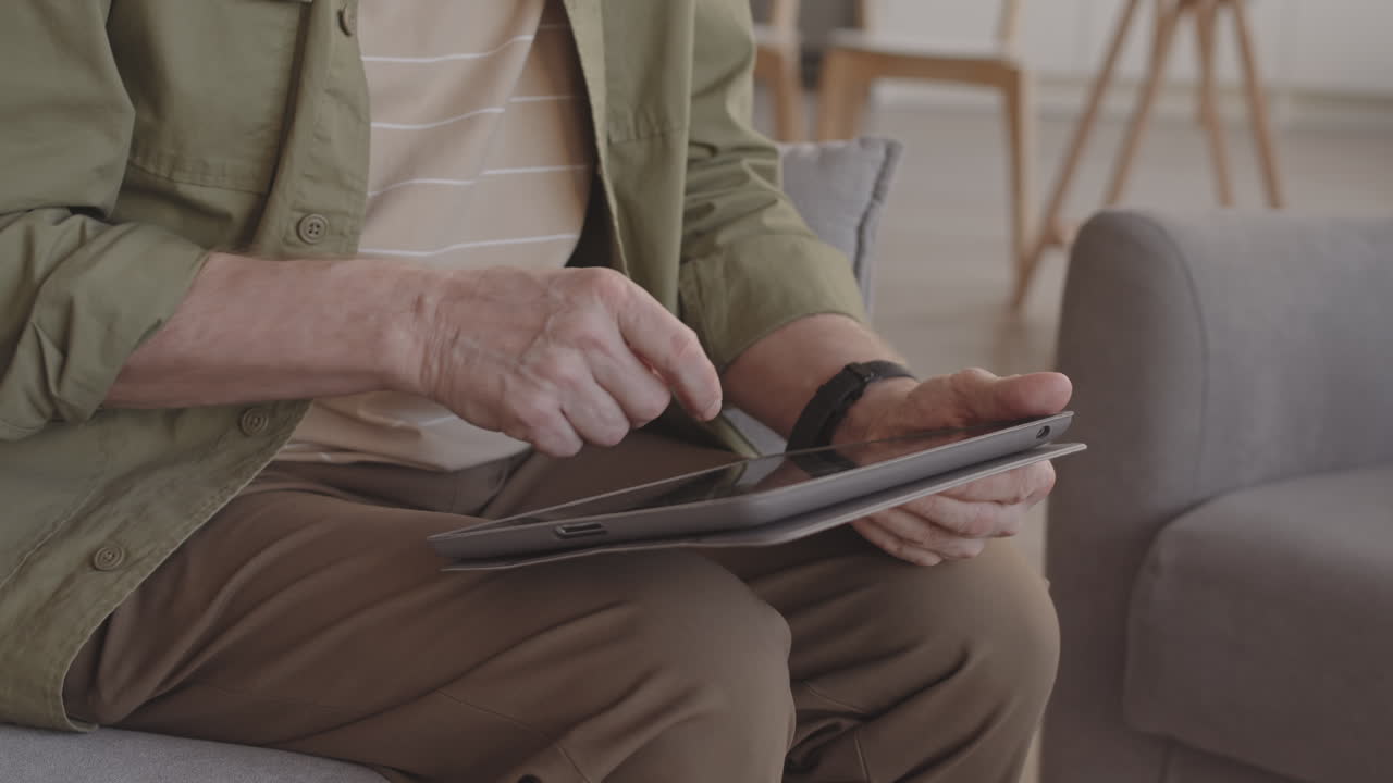 Tilting up of older Caucasian man wearing glasses, scrolling on screen of tablet computer, sitting in armchair in living room