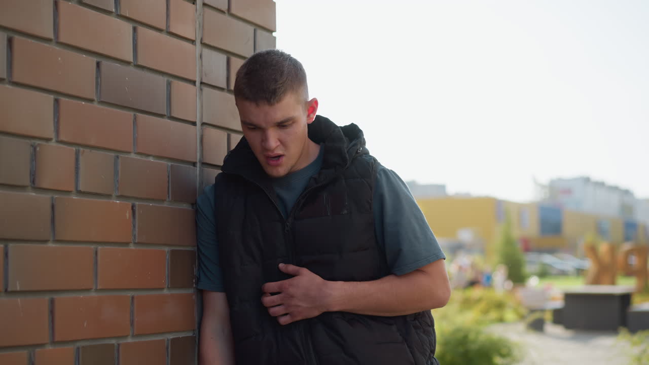 white man in black puffer vest leaning on brick wall in urban environment exhaling dense vape smoke and coughing into hand after long inhale of portable vape device