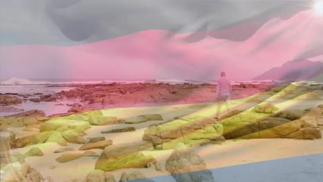 Animation-of-flag-of-germany-over-senior-caucasian-man-walking-beside-rocks-on-beach-against-sky