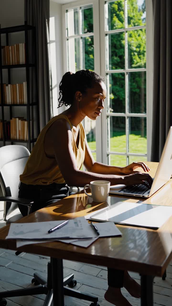 A woman works on a laptop in a sunlit room, captured from a side angle