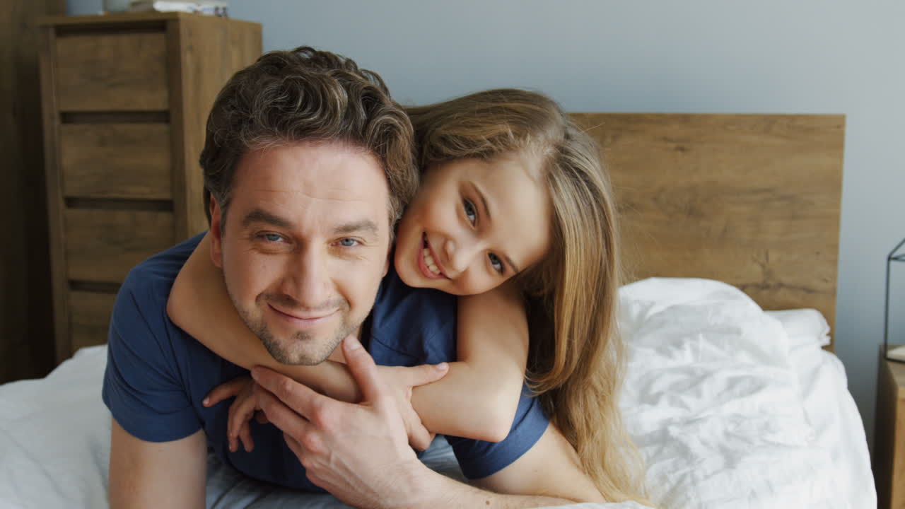 Close-up view of happy father and his blonde daughter hugging and lying on the bed while looking at camera