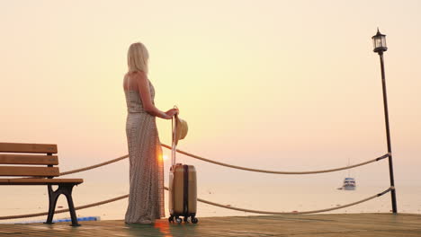 a young girl in a summer sundress expects to settle in a resort hotel stands with a suitcase on the