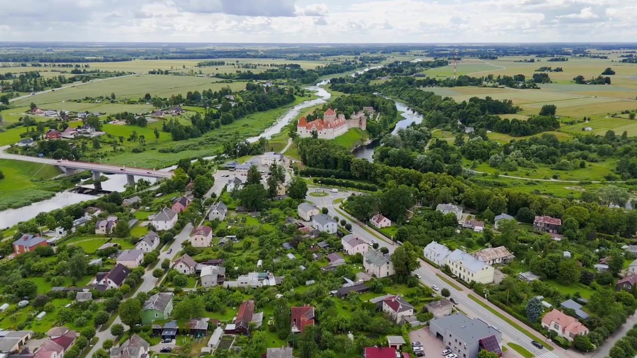 Dronekuva Bauskasta, Latviasta, näyttäen Bauskan linnan kahden joen välissä, asuinkatuja, siltoja, vihreää maaseutua ja viljelysmaiden horisontin.