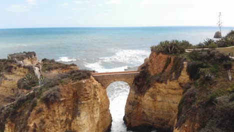 Stone-Bridge-connecting-eroded-cliffs-in-Student-Beach-In-Lagos,-Algarve,-Portugal---Aerial-dronie-ascending-wide-view-shot