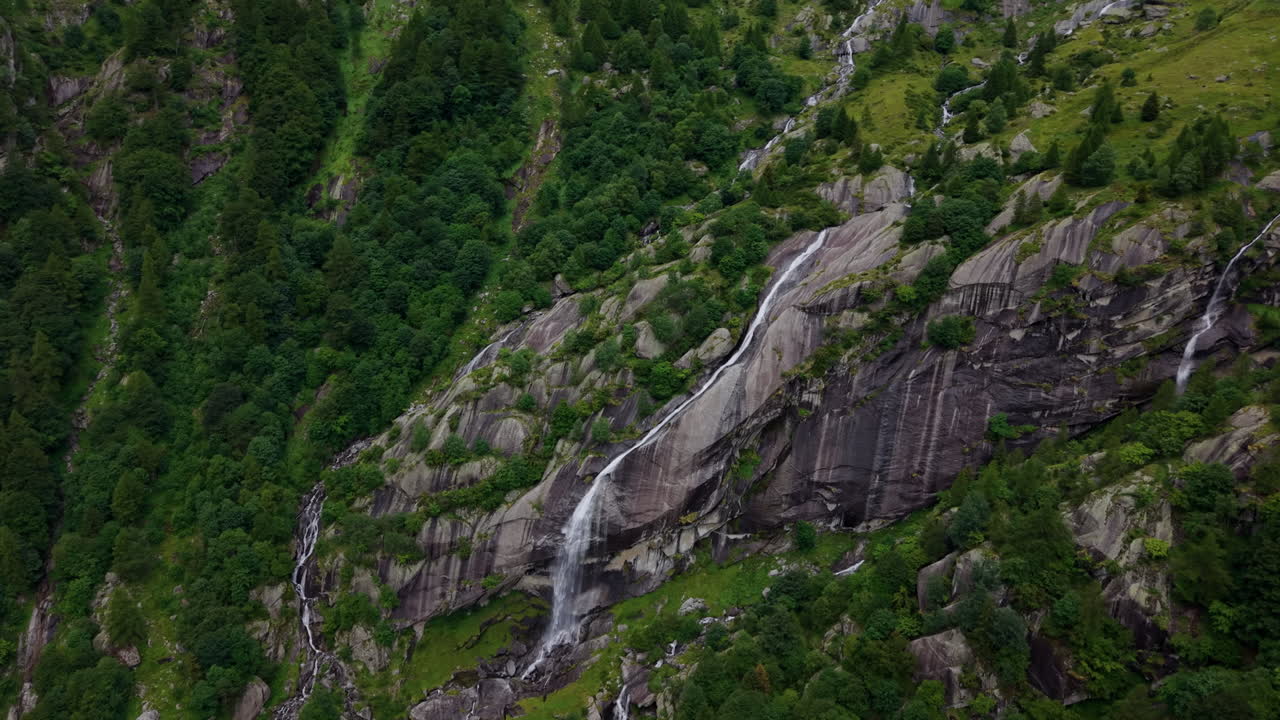 Aerial drone shot advancing toward a long waterfall on a mountain slope, performing a tilt down over the rocky terrain and forest in the Swiss Alps