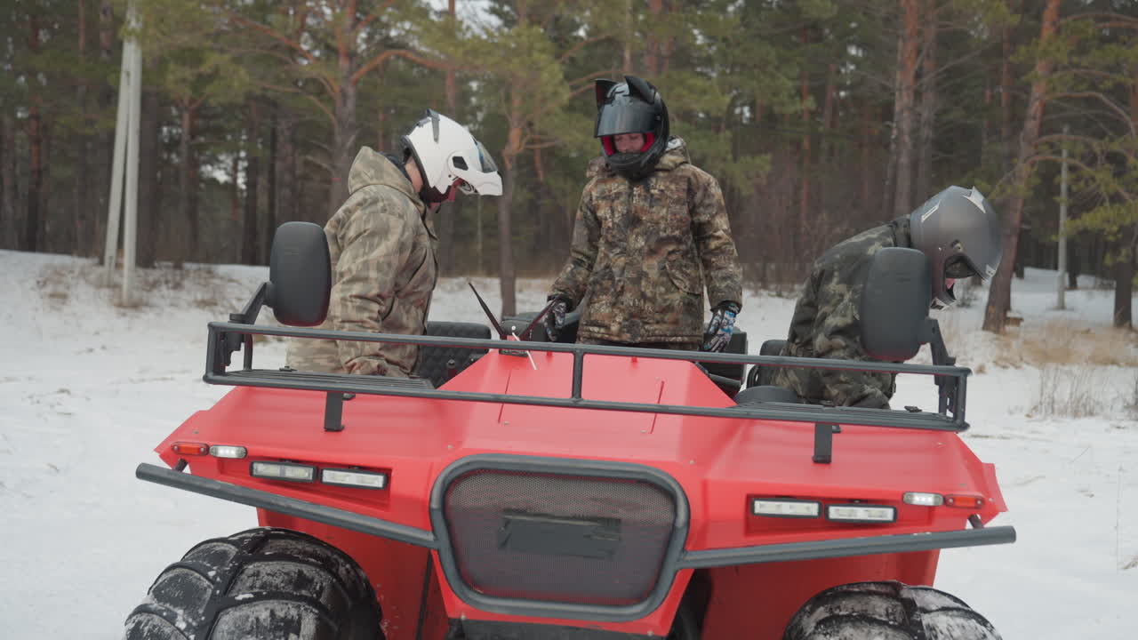 Un istruttore assiste i piloti a salire su un quad rosso nella neve. L'equipaggio concentrato, in mimetica e con i caschi, esegue controlli di sicurezza, sale sul veicolo, ispeziona pneumatici e motore e discute il percorso in una pineta, in un'atmosfera tesa.