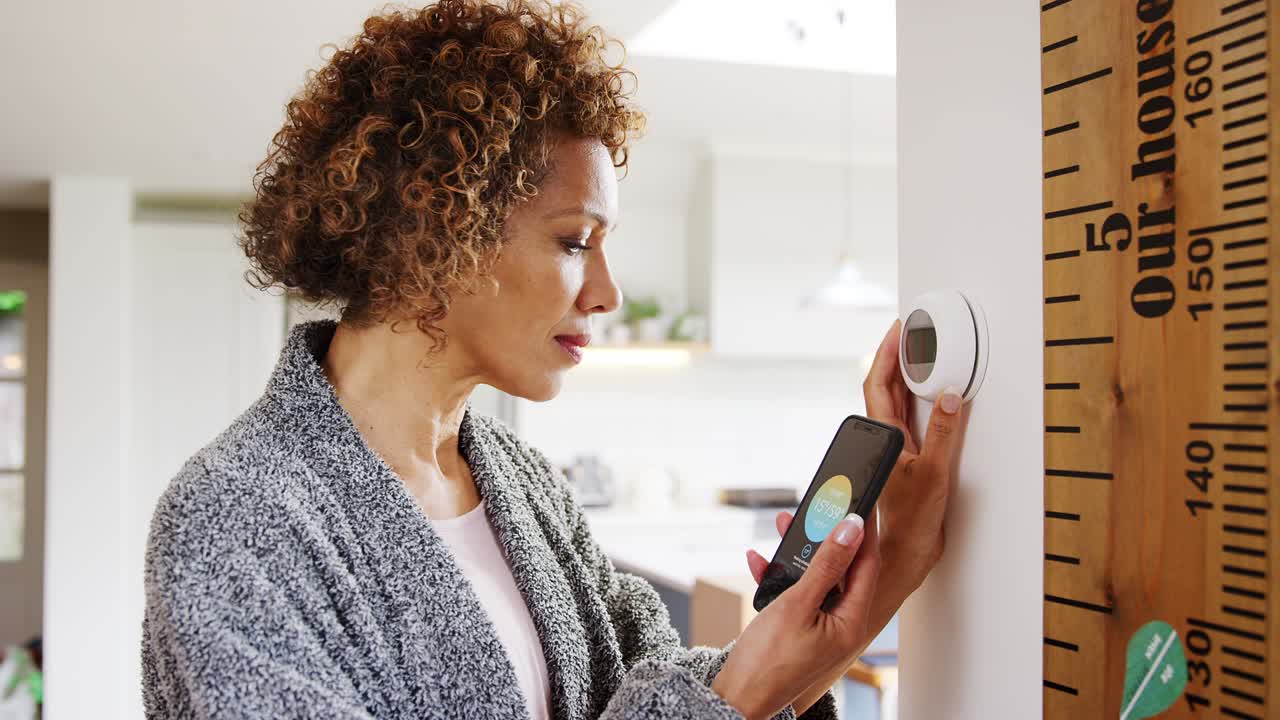 Mature woman using app on phone to control digital central heating thermostat at home with gay partner in background - shot in slow motion