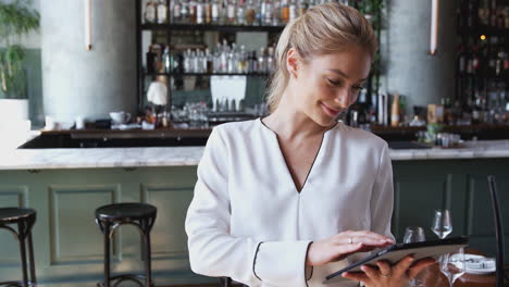 Female-Owner-Of-Restaurant-Bar-Standing-At-Counter-Using-Digital-Tablet