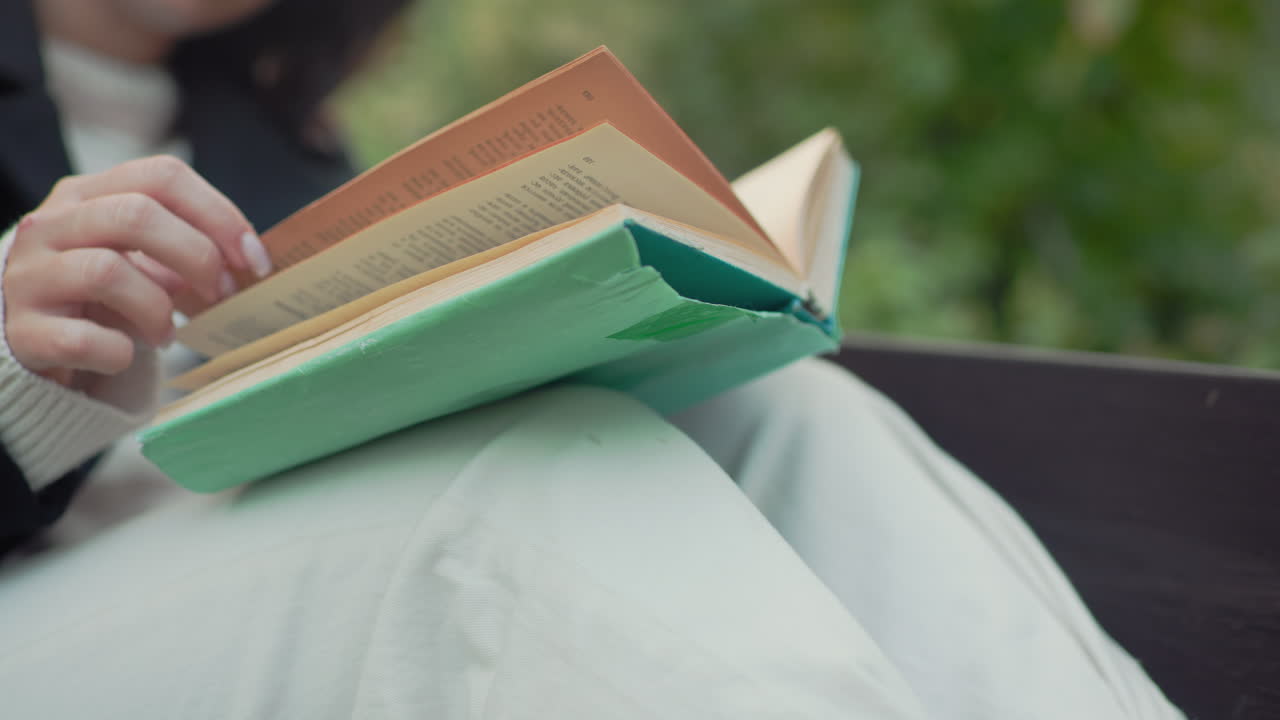 Lower view of relaxed lady seated on bench with legs up, holding worn green novel on lap, softly flipping pages, adjusting hair between lines, paper cup on ground, calm autumn park background