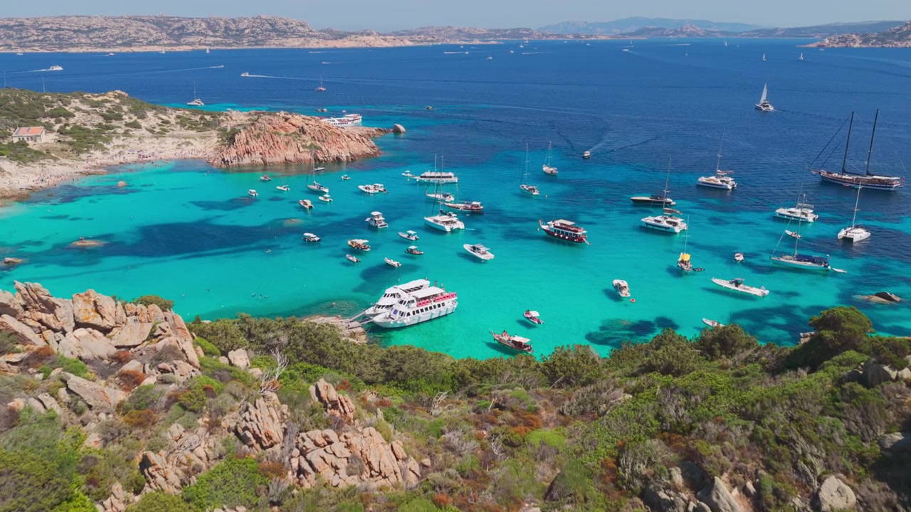 Vue aérienne épique survolant des falaises de granit pour révéler des yachts à voile de luxe dans l'eau cristalline d'un port naturel protégé sur l'île de Spargi dans l'archipel de la Maddalena en Sardaigne, Italie.