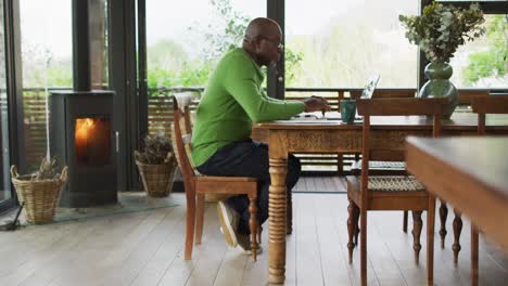 African-american-senior-man-sitting-at-dining-table-working,-using-laptop