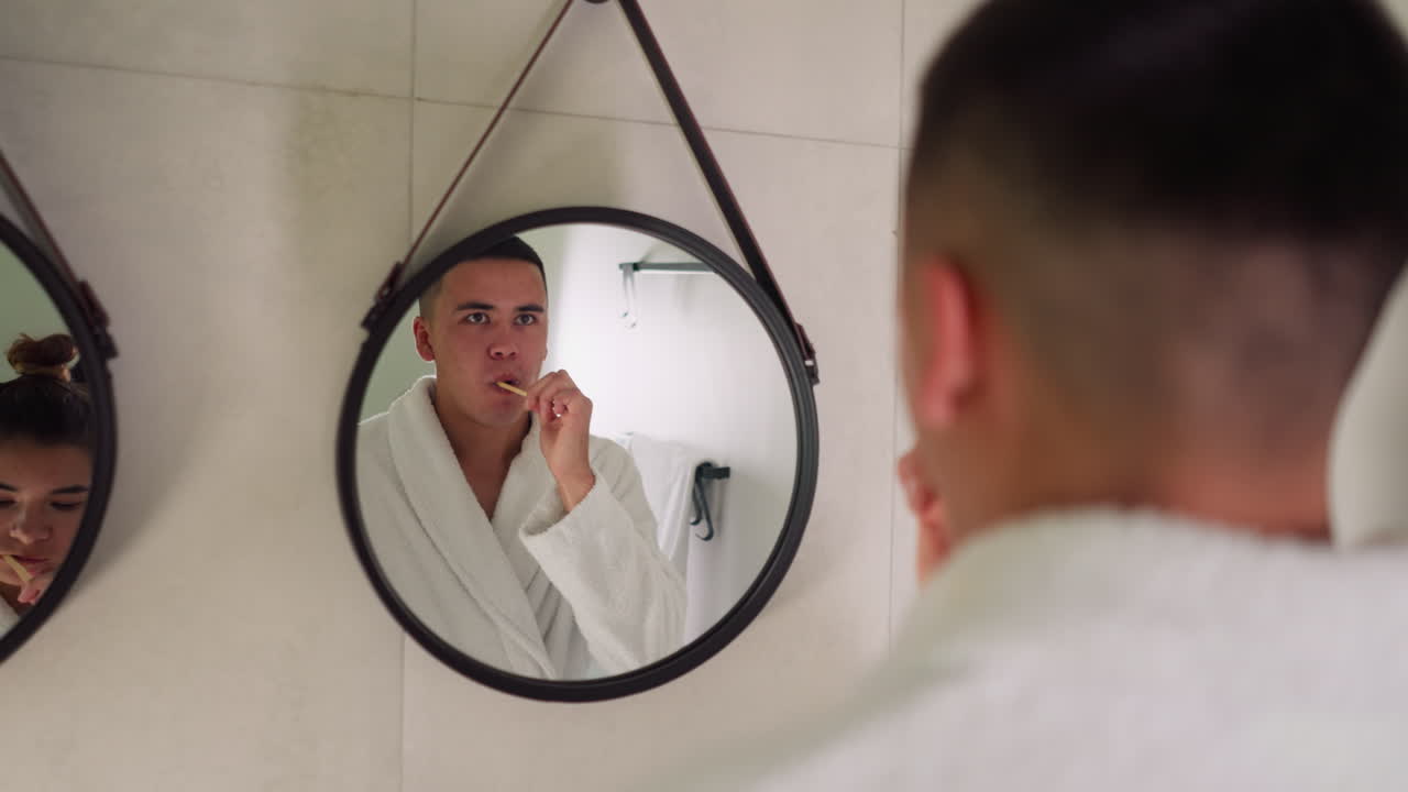 el marido se cepilla los dientes con el espejo en casa. el hombre en bata de baño de terry realiza procedimientos de cuidado diario para la salud dental de pie cerca de la esposa en el baño