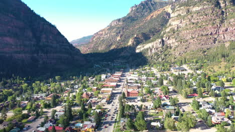 aerial drone footage of ouray colorado mountain town, cars driving through city and houses surrounded by rocky mountain cliffs and pine tree forest