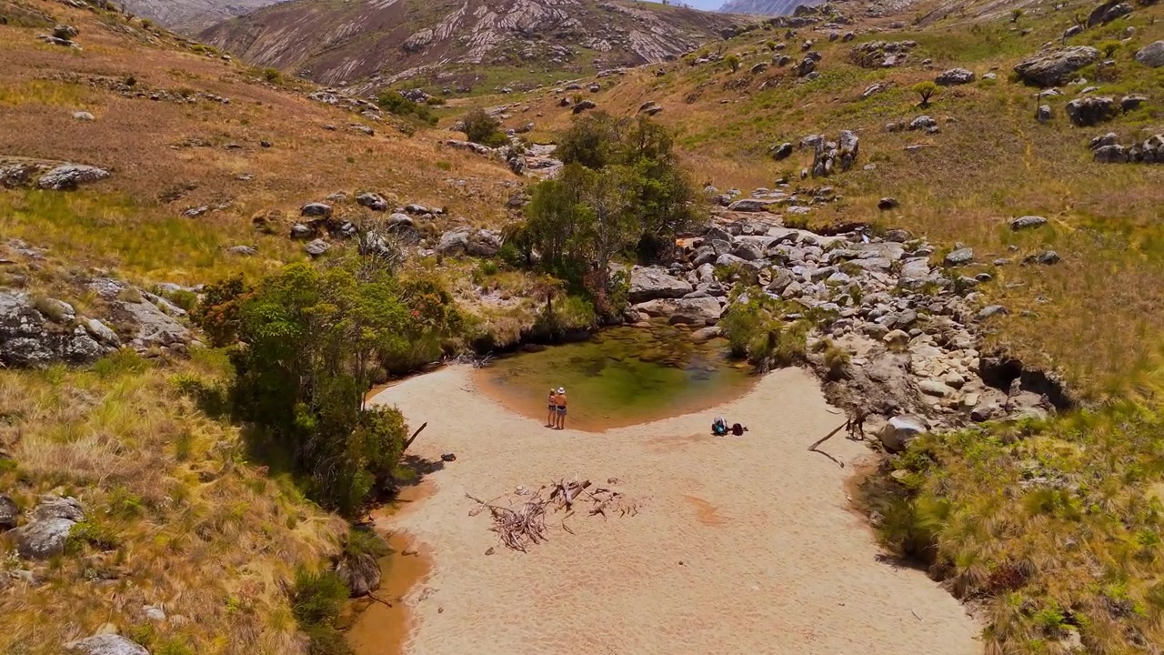 Un drone si avvicina a una spiaggia fluviale sabbiosa dove alcune persone sostano accanto a una piscina naturale, circondata da rocce e praterie nel massiccio del Mulanje, in Malawi