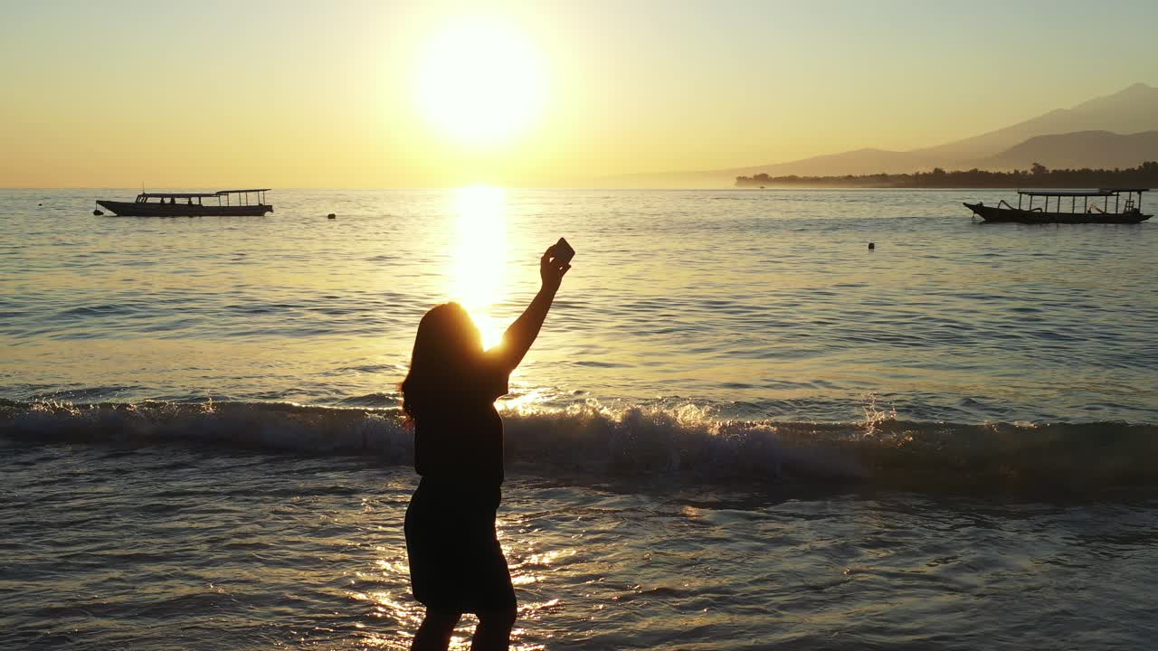 silueta de una mujer joven tomando fotos de selfie en una playa tranquila a la hora dorada del atardecer con el cielo amarillo reflejado en la superficie del mar