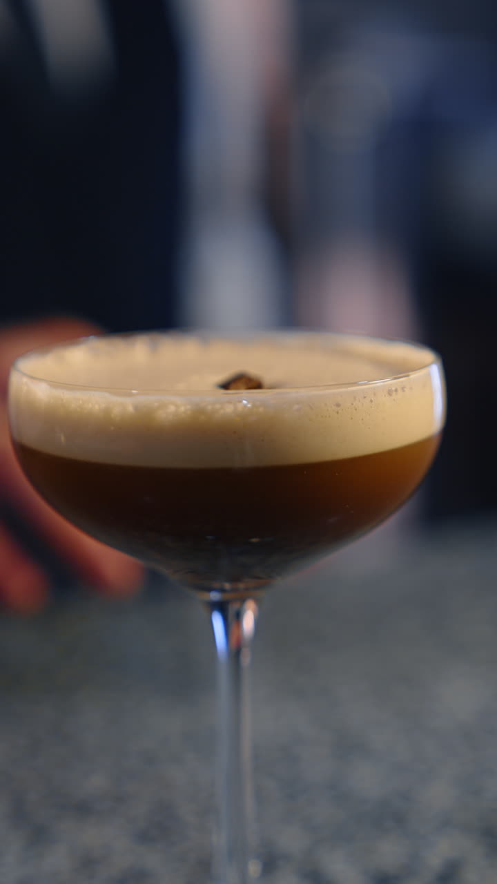 A coupe glass with a brown drink and creamy foam. Bartender uses the metal tongs to put a coffee grain into the beverage. Close up. Vertical video.