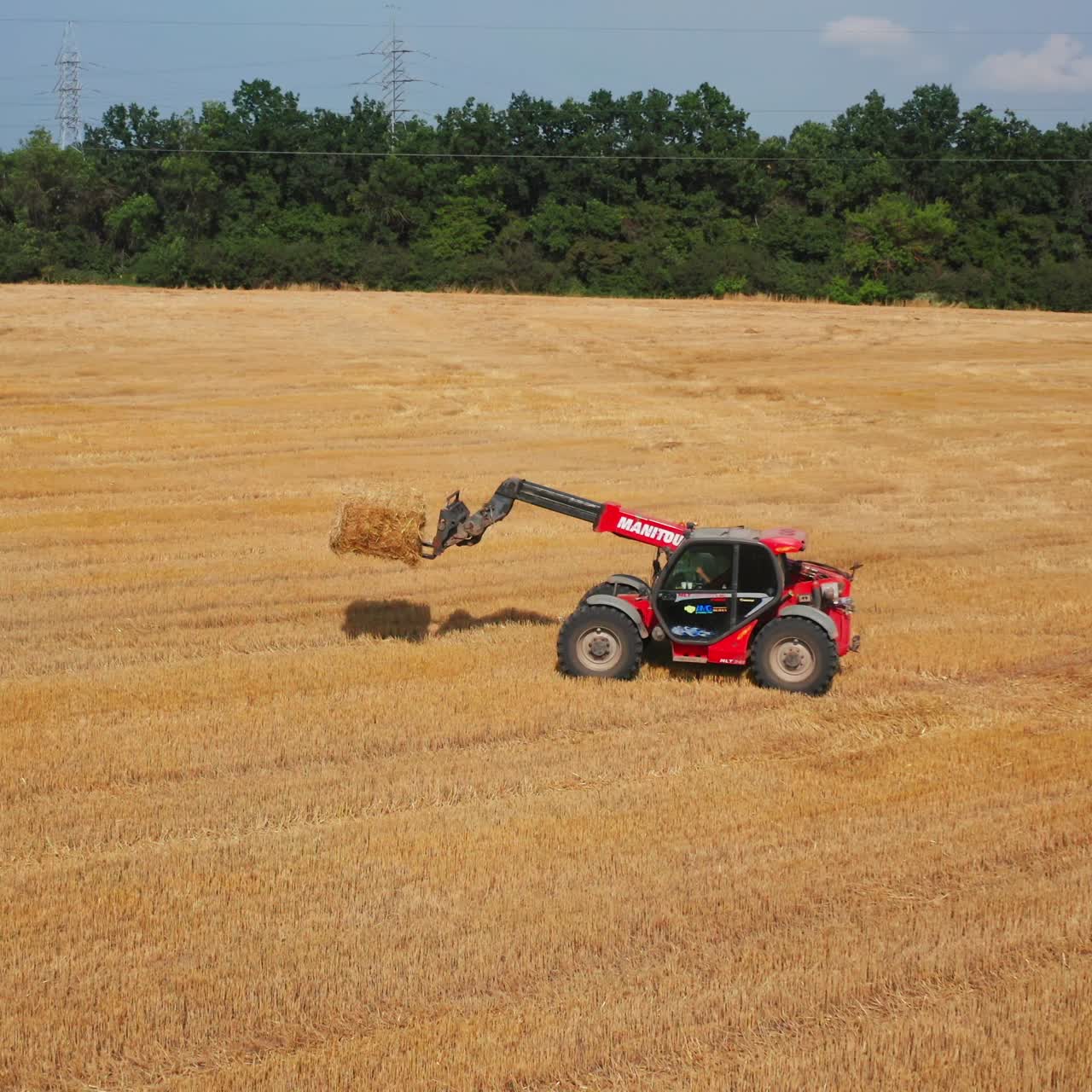 Tractor goes by the field followed by the loader carrying hay bale. Agricultural technique picking straw after harvesting wheat grain