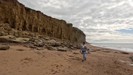 female walking along a jurassic coast beach alone on a cloudy morning captured in slow motion. cameraslowly pans to the left as the women walks to the left across the shot.