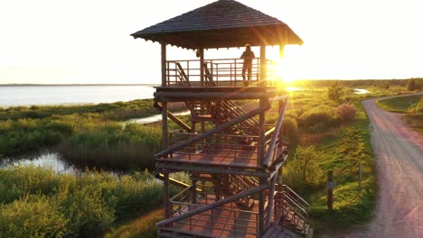 Girl-observing-golden-sunrise-from-top-platform-wetland-birding-hide-observation-hunting-tower-aerial-push-in