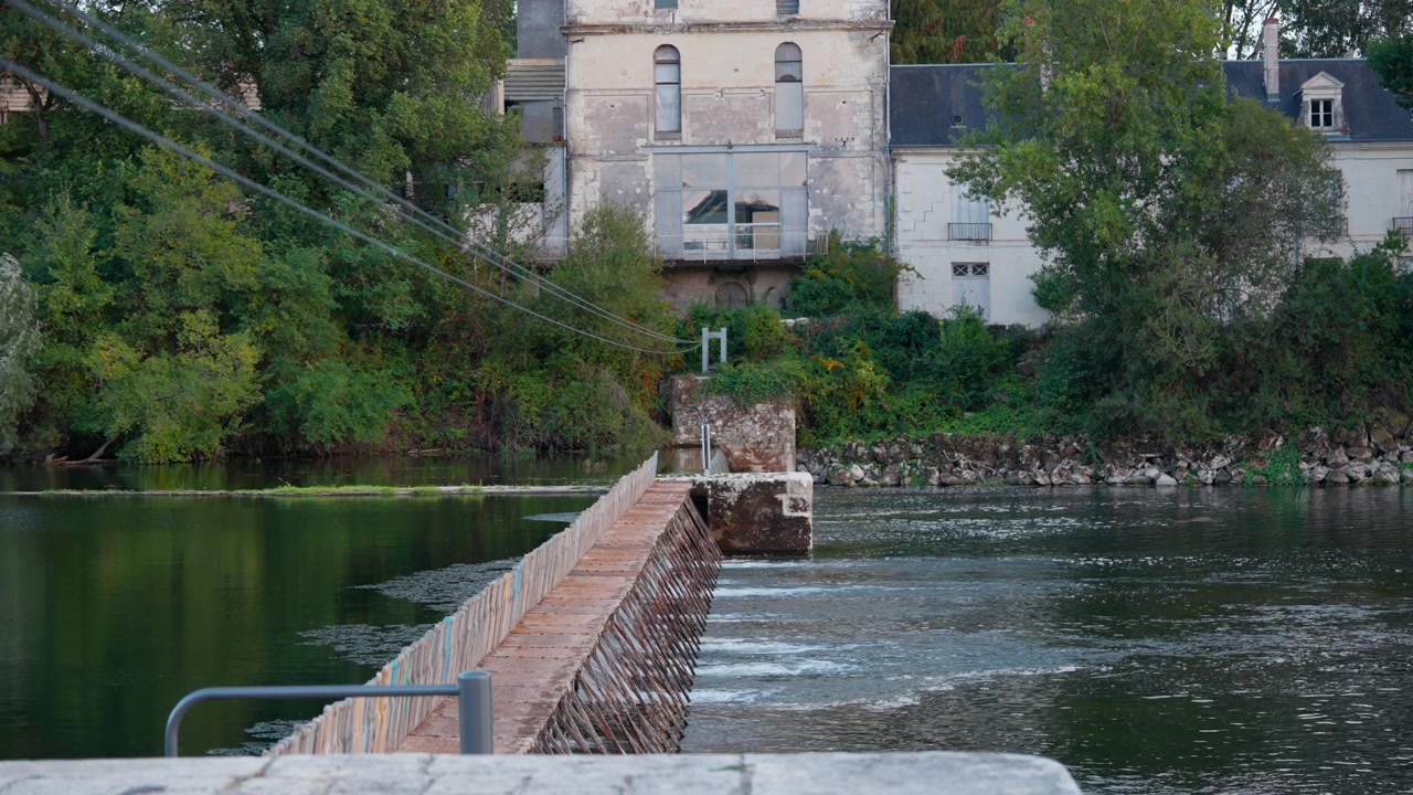 Storica diga ad aghi che attraversa un fiume calmo con vecchi edifici e vegetazione rigogliosa nelle vicinanze