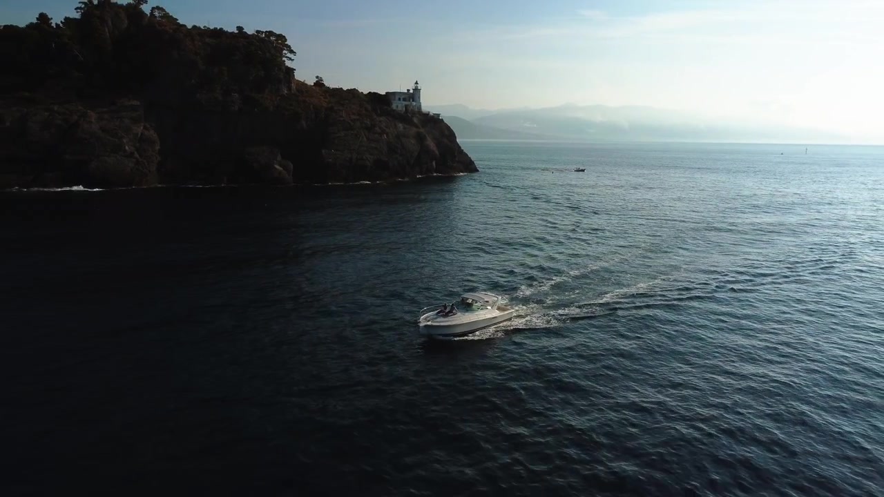 Vista aerea che si allontana da un motoscafo di fronte al Faro di Portofino, giornata estiva in Italia