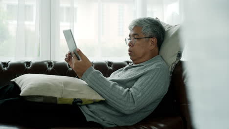 active senior man relaxing at home working, wearing bluetooth earbud enjoy listening to music from a mobile phone sitting comfortably on a sofa