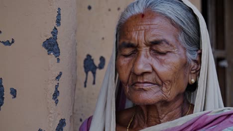 retrato de una anciana serena con cabello gris y un pañuelo tradicional, parpadeando y moviendo sutilmente su cabeza contra una pared desgastada